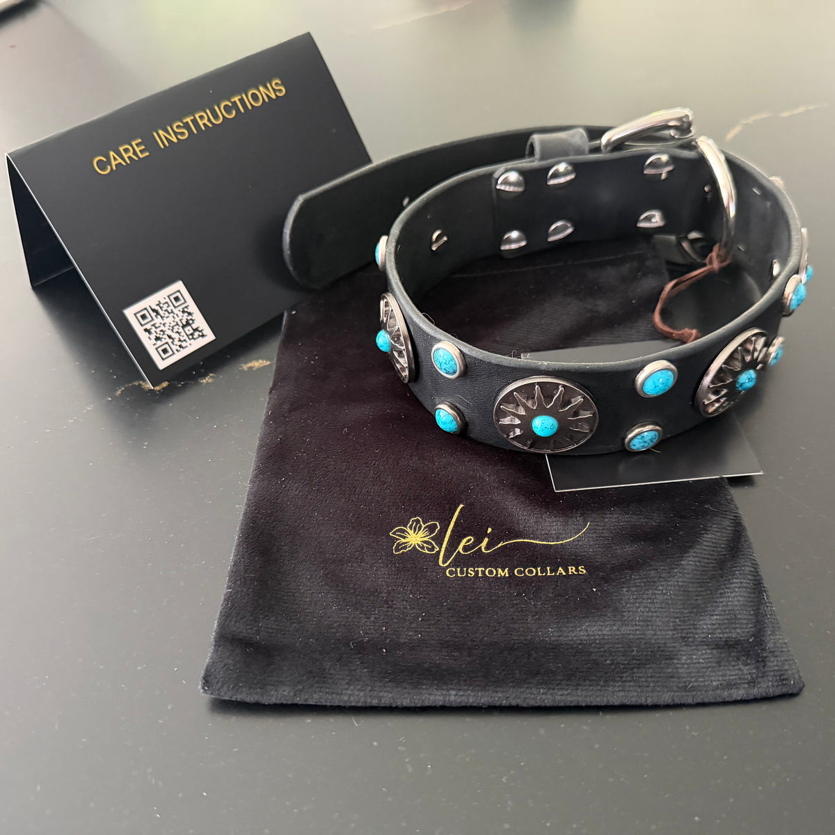 A black leather dog collar with silver-tone conchos and turquoise accents, displayed on a black velvet pouch branded 'Lei Custom Collars' with a care instructions card beside it.