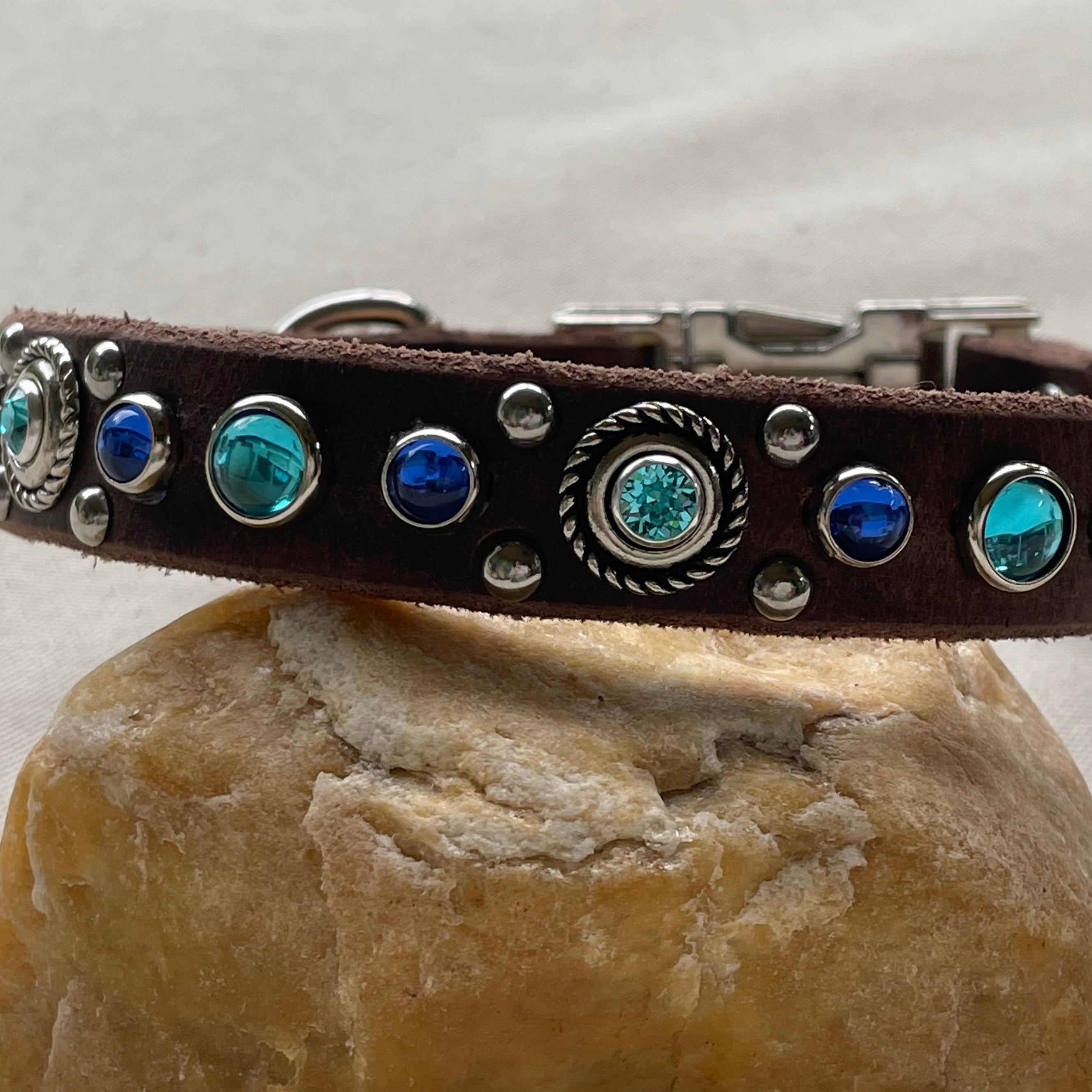 Brown leather dog collar with blue and silver embellishments on a stone surface