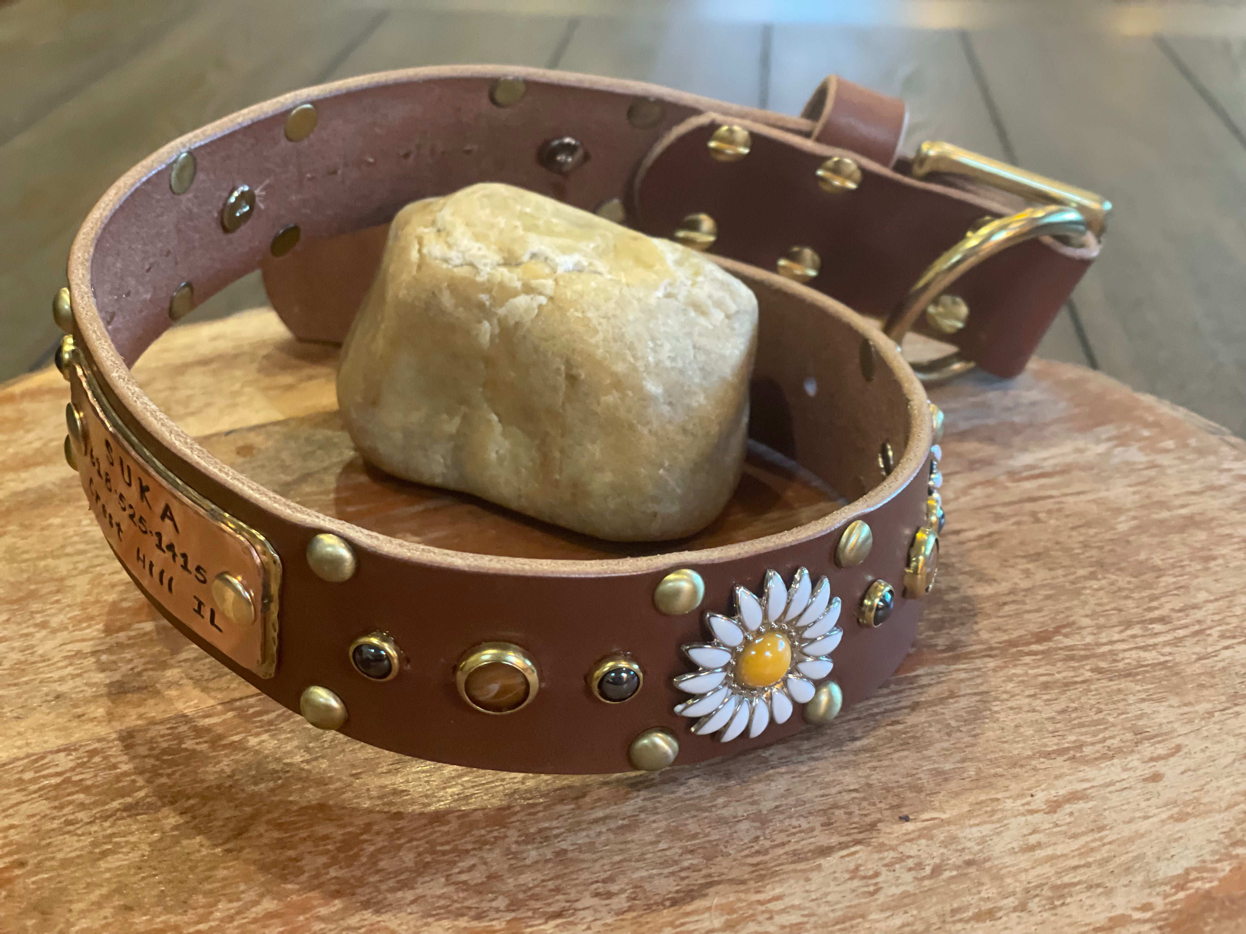 Custom brown leather dog collar with engraved brass name plate, daisy flower concho, hand-set stones, and solid brass buckle, artisan pet collar displayed on wood with natural stone.