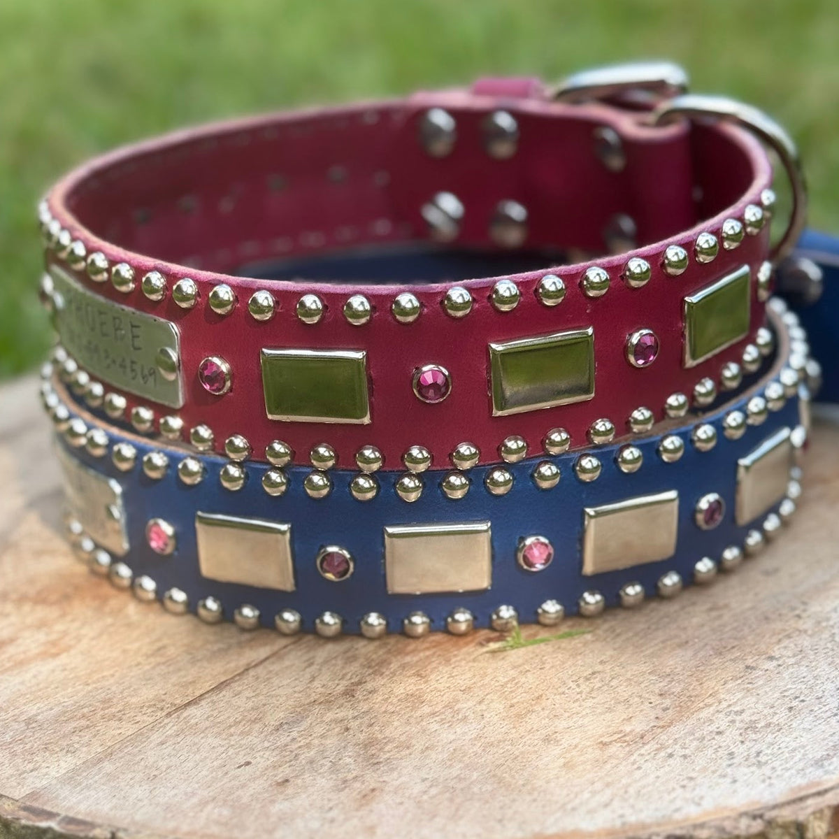 Two decorative leather dog collars stacked on a round wooden log outdoors. The top collar is burgundy with silver studs and rectangular metal plates, and the bottom collar is navy blue with matching silver studs and plates.