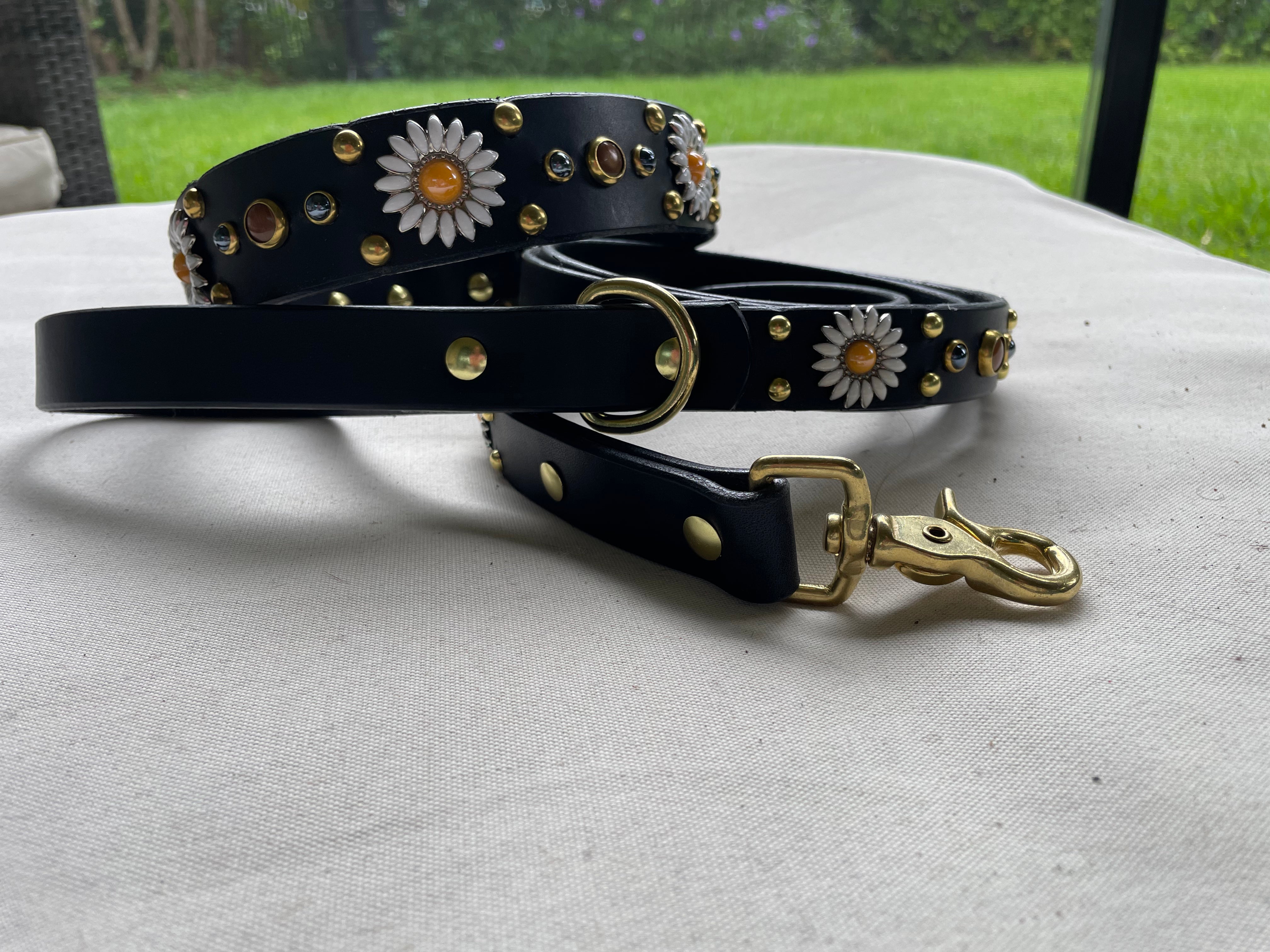 Black leather dog collar with matching studded leather leash, brass swivel clip and D-ring, daisy concho detailing, and handcrafted luxury pet hardware set photographed outdoors.