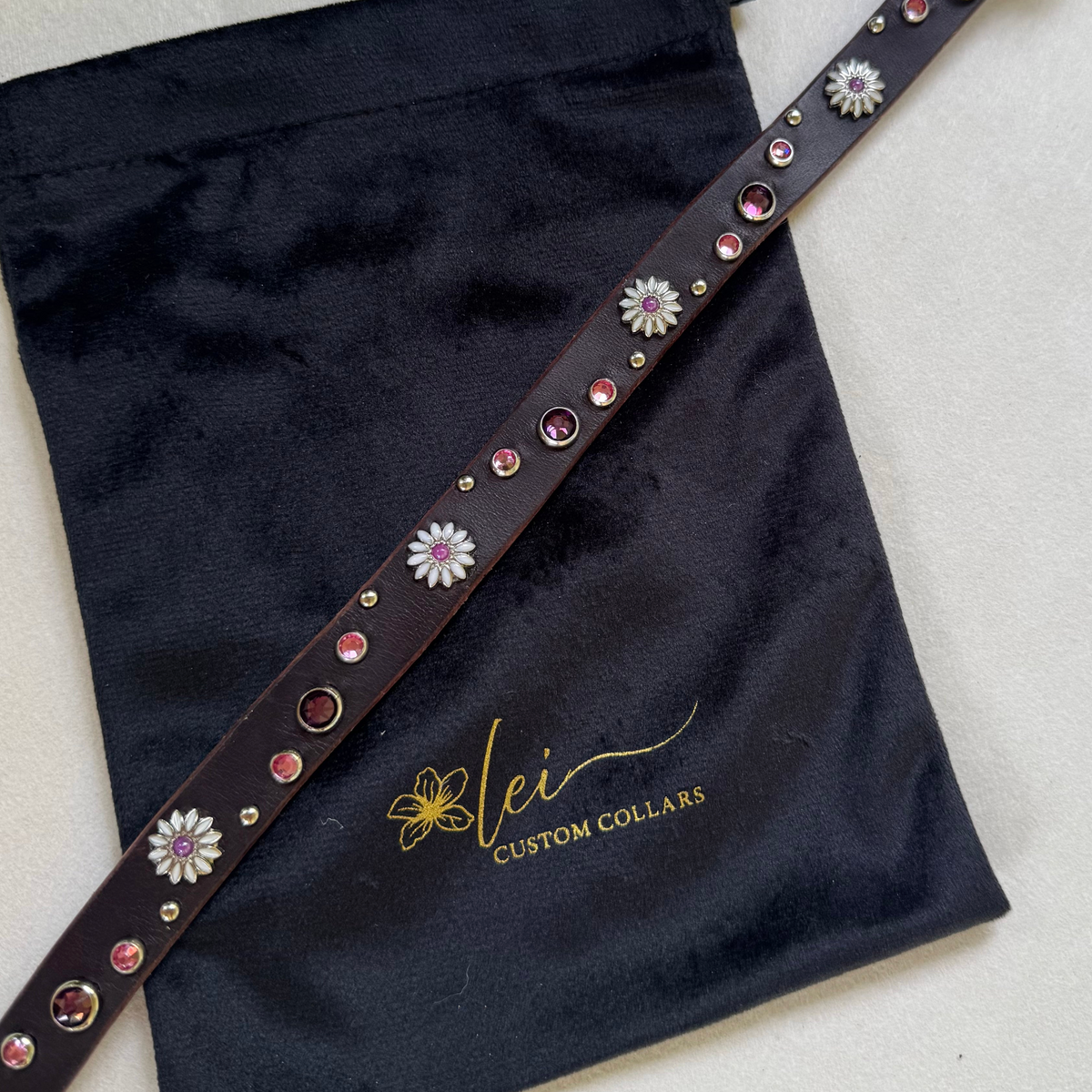 A dark navy drawstring bag with a decorative embroidered strap featuring white floral motifs and colorful beads, shown on a light surface. The bag has 'Lei Custom Collar' embroidered in gold on the front.