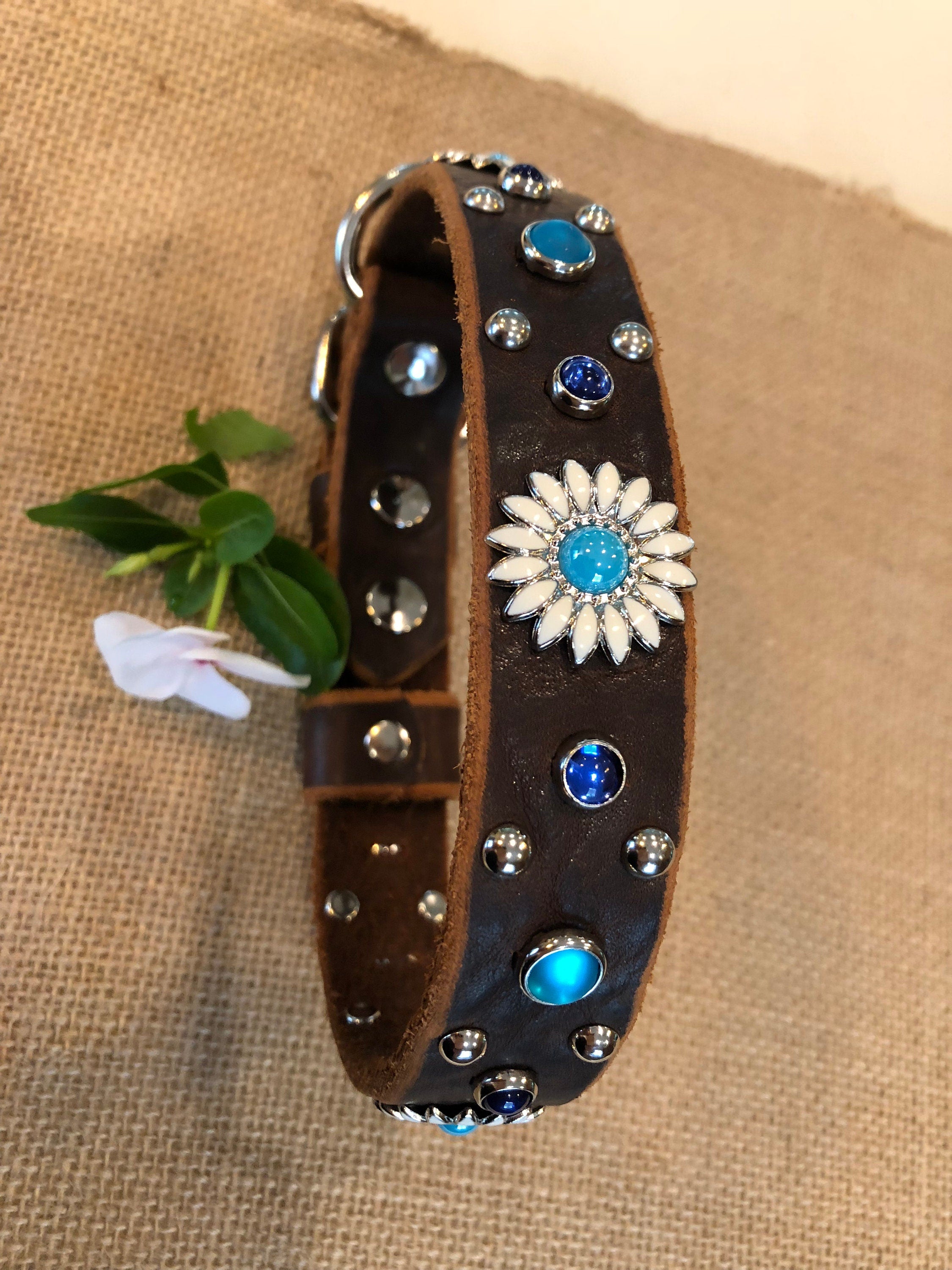 Close-up of a brown leather dog collar featuring a daisy-style concho centerpiece, turquoise and deep blue crystal accents, and polished metal studs, styled on textured fabric with a white flower for an elegant artisan finish.