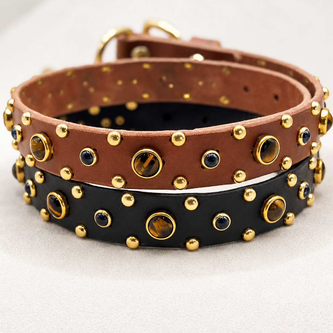 Two studded leather collars stacked on a light gray surface, one brown and one black, both decorated with gold-tone metal studs, round tiger's eye gems, and small black oval stones.