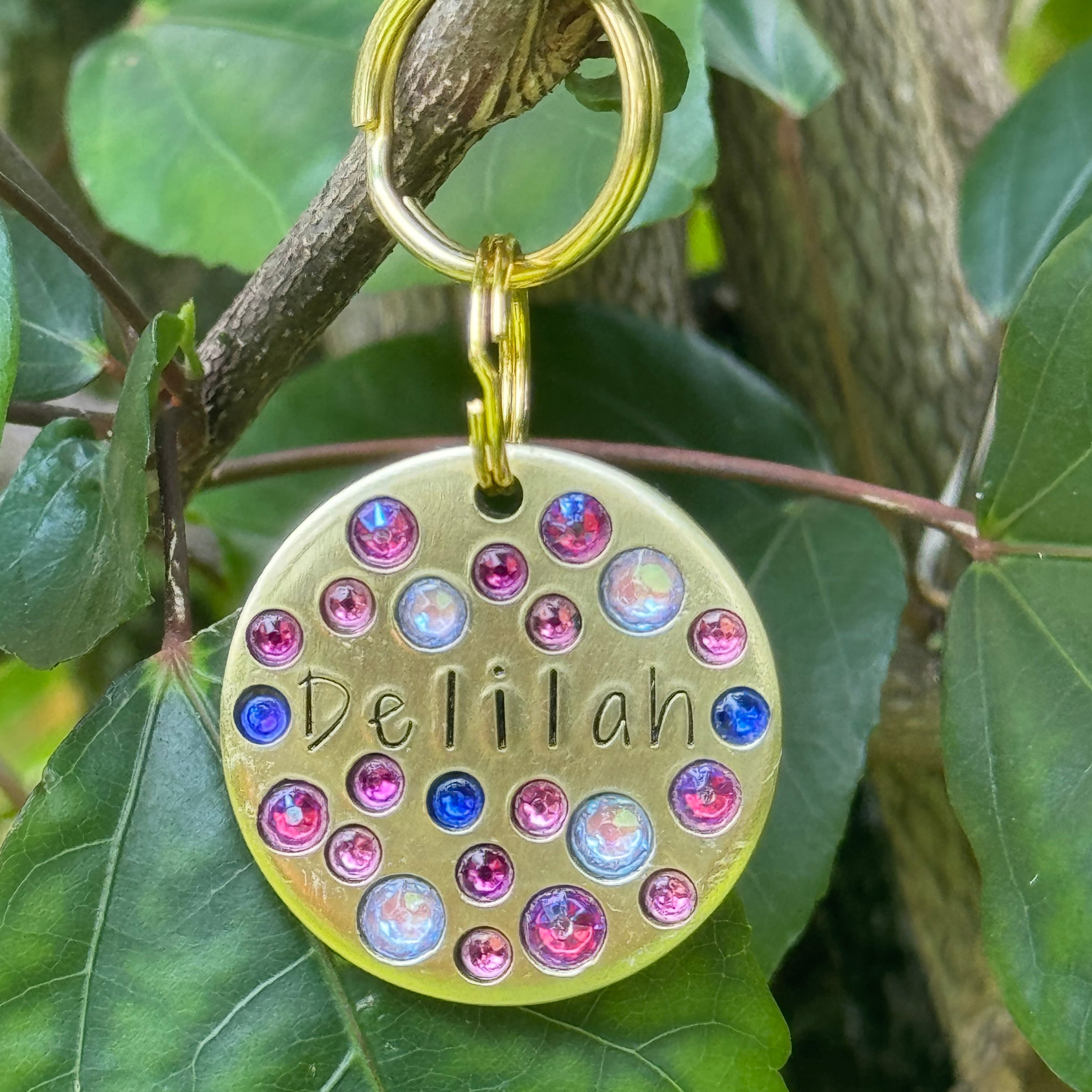 Gold dog tag with colorful rhinestones and 'Delilah' engraved, hanging on green leaves.