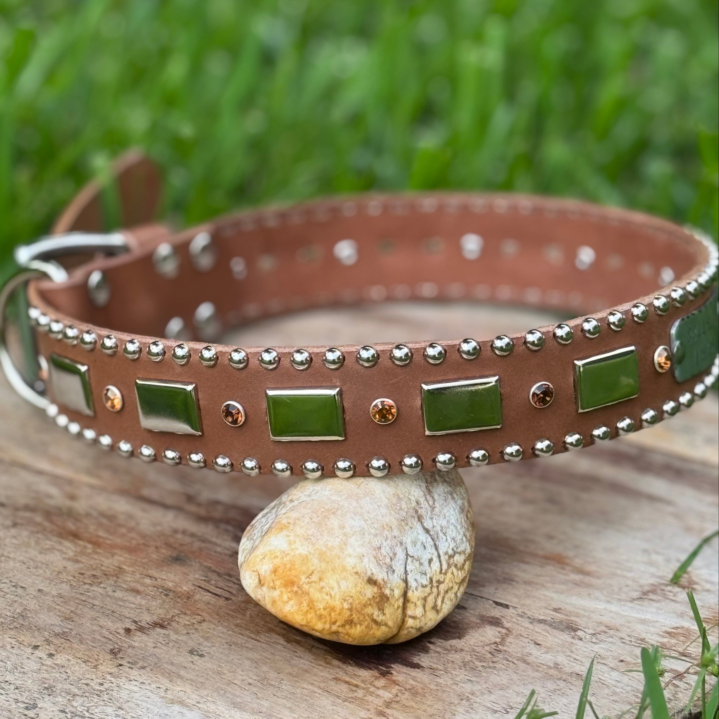 Brown dog collar with green accents and silver studs on a wooden log with grass in the background