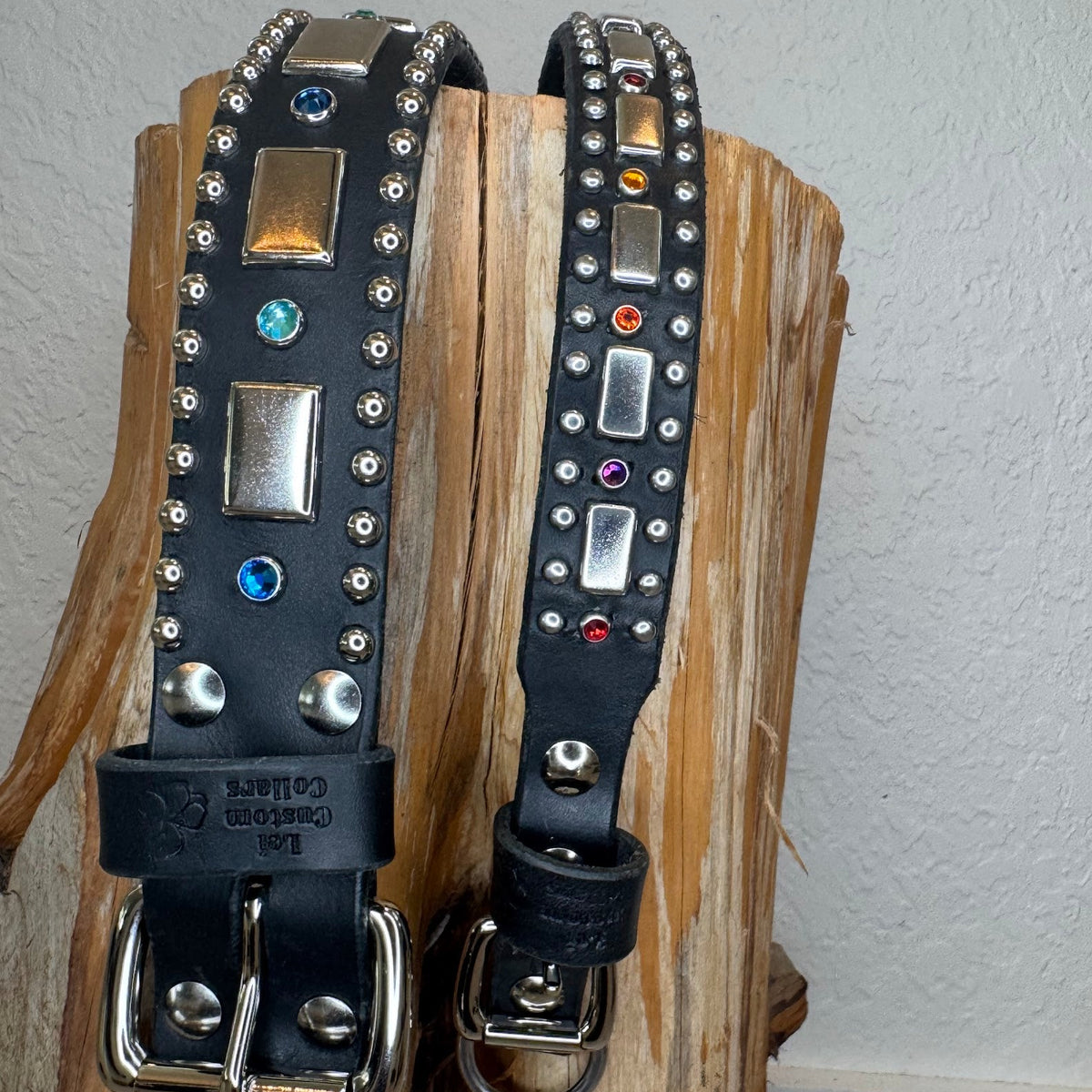 Two black leather dog collars 1.5" wide and 1" wide with silver-tone hardware and colorful rhinestone accents, displayed on a rustic wood block against a white wall.