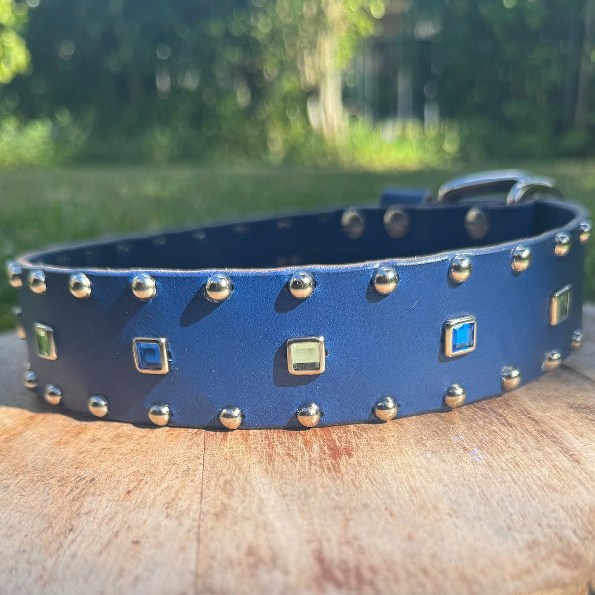 A blue leather dog collar with gold-tone studs and decorative square accents, displayed on a rustic wood log outdoors with green grass and trees in the background.