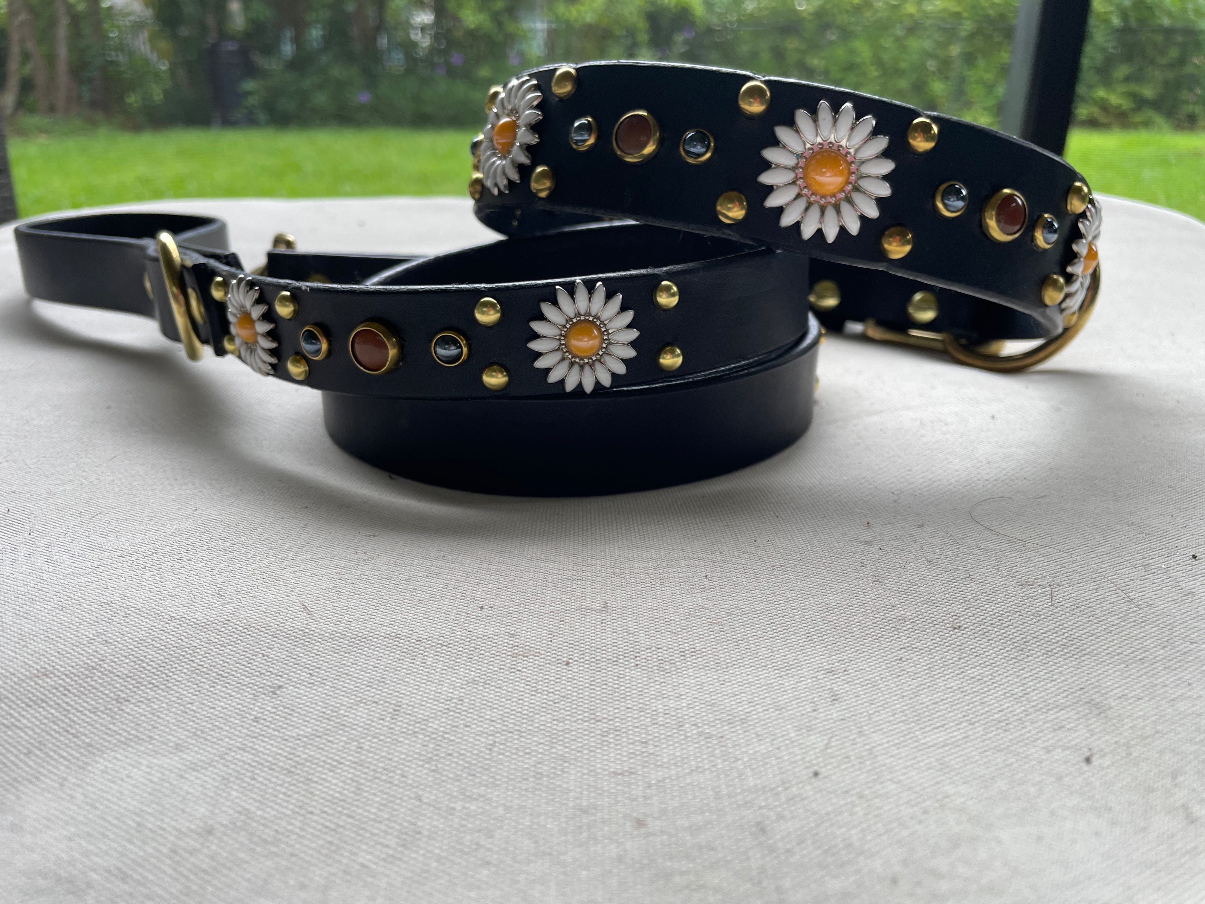 Black handcrafted leather dog collar paired with matching leather leash, featuring brass hardware, daisy floral conchos, and stone stud accents, luxury pet set photographed outdoors.