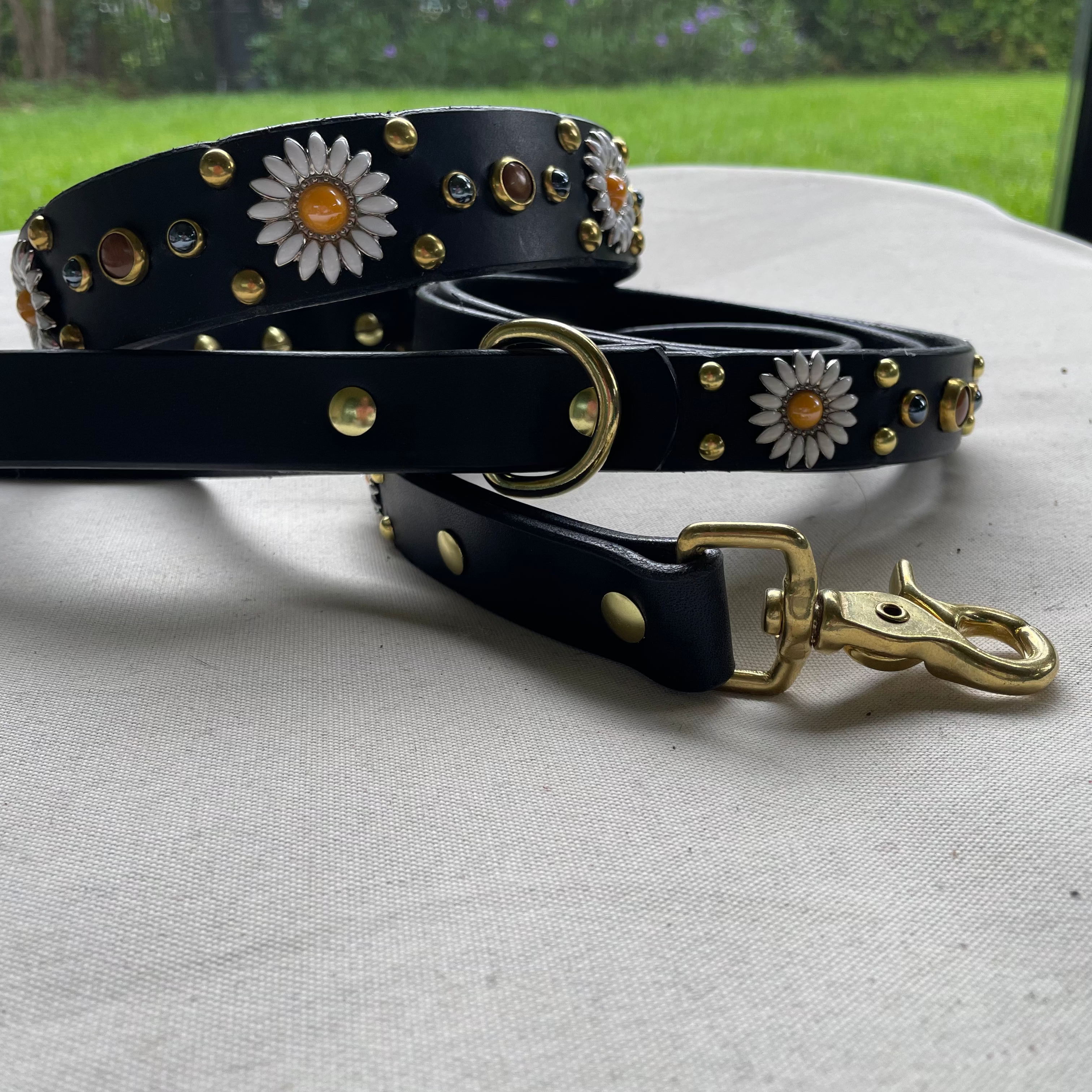 Black leather dog collar with matching studded leather leash, brass swivel clip and D-ring, daisy concho detailing, and handcrafted luxury pet hardware set photographed outdoors.