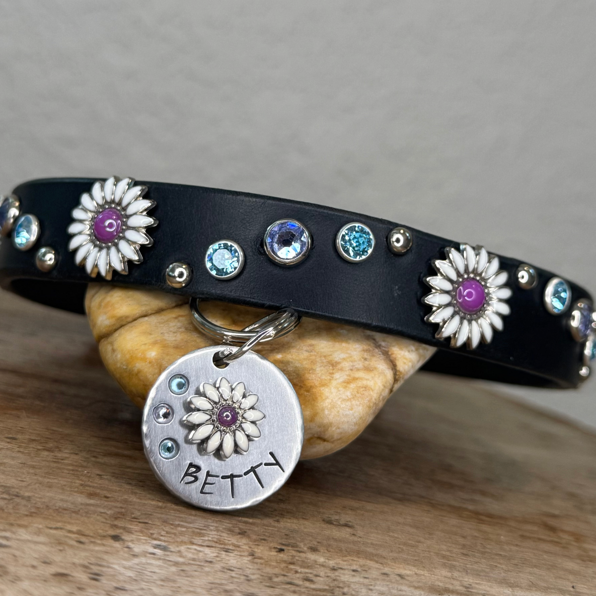 A black leather collar with silver floral studs, blue and purple rhinestones, and a silver 'BETTY' charm, displayed on a wooden surface with a stone. The image is shown within a Shopify website editing interface on the right side.