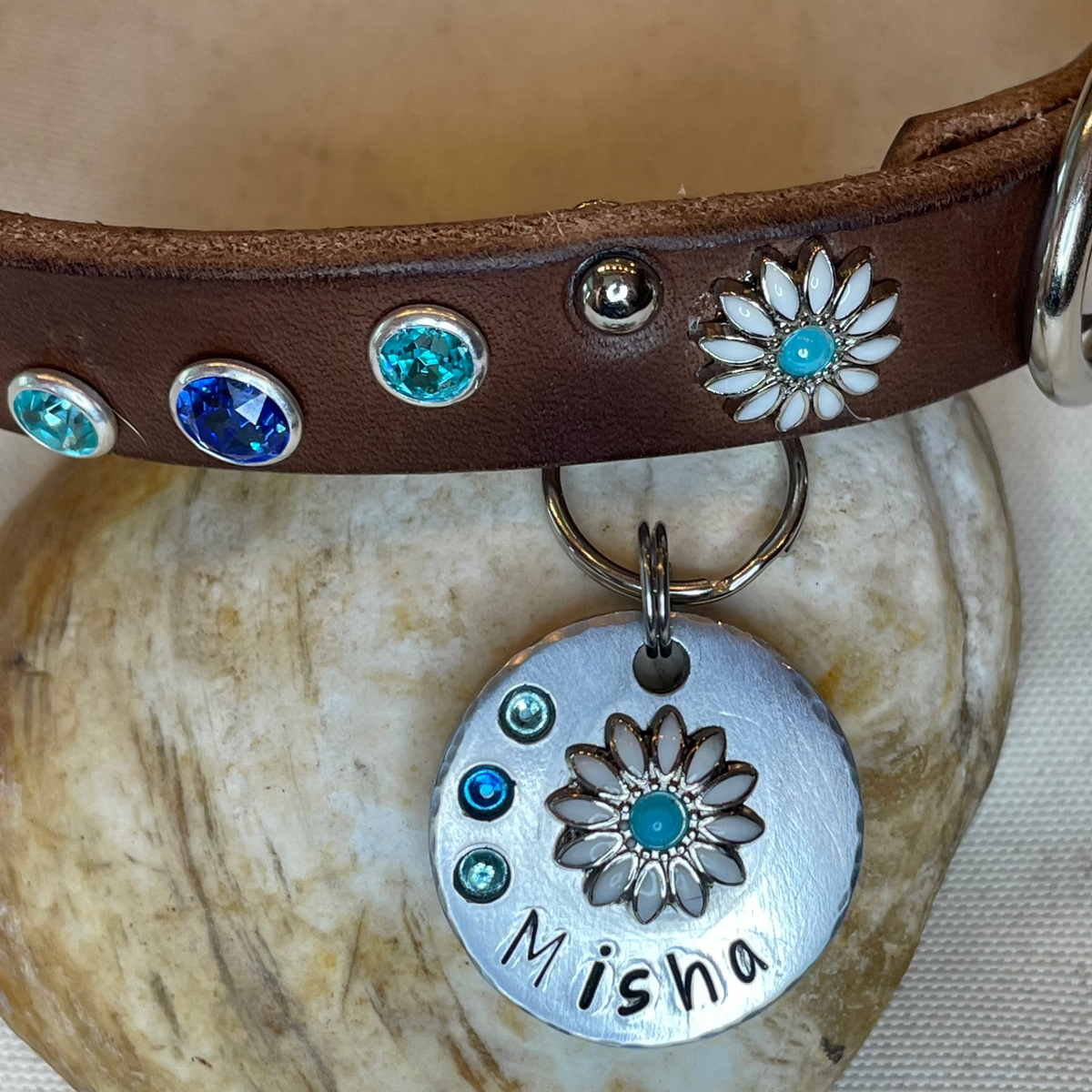 A custom engraved dog collar shown on a shell-shaped surface, featuring a brown leather collar with turquoise and silver floral studs and a round silver tag engraved with the name 'Misha' and a flower design. The image is a screenshot of a product customization page with text fields and editing tools visible on the right.