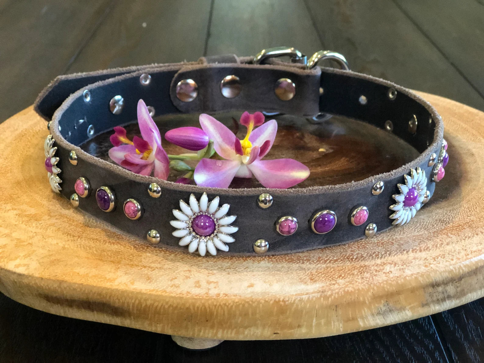Dark leather dog collar with white daisy conchos, purple stone accents, and silver rivet detailing, handcrafted custom pet collar styled with pink flowers on a wooden stand.