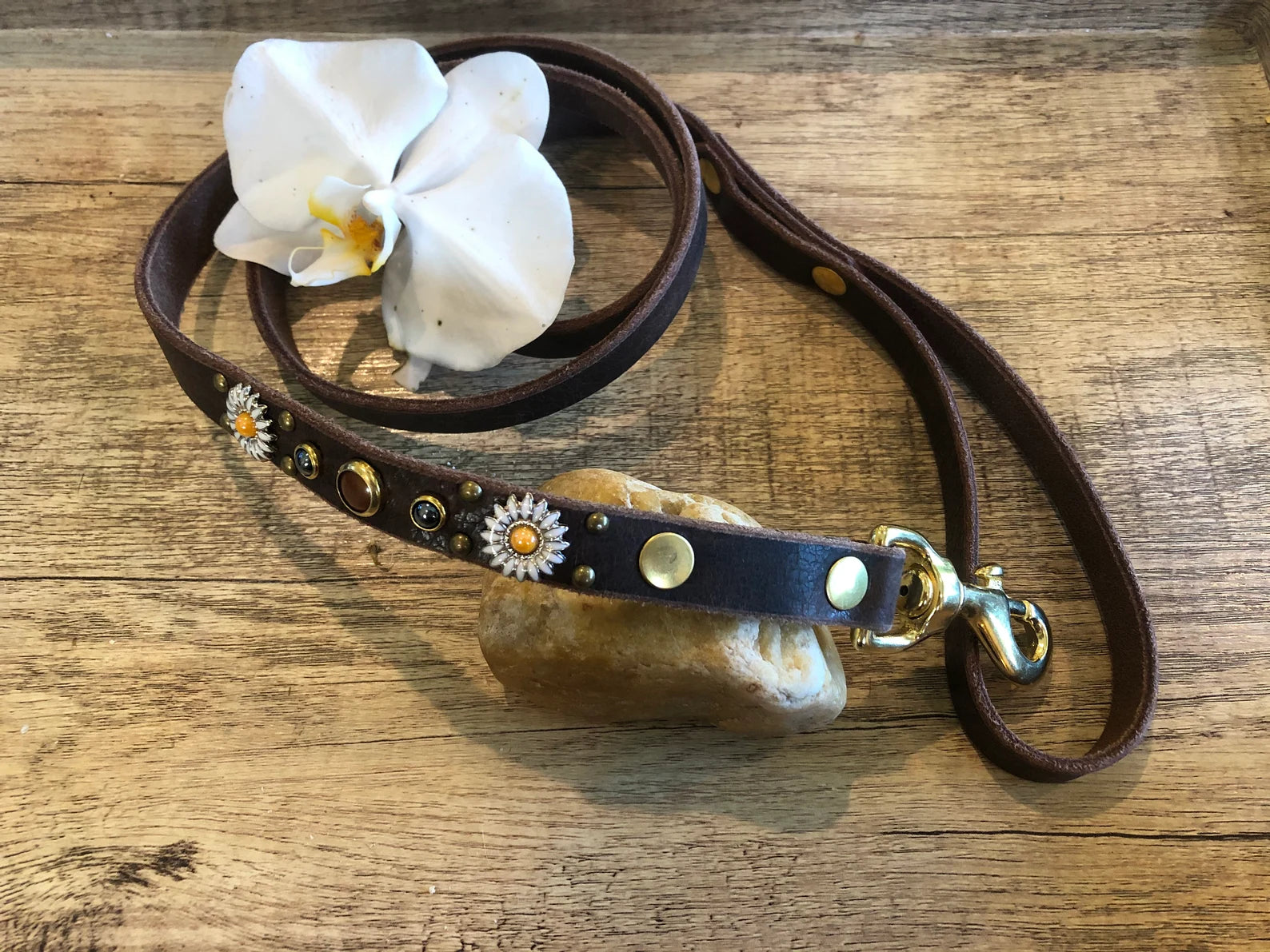 Handcrafted luxury leather dog leash in rich brown, featuring gold hardware and floral crystal concho details, styled with a white orchid on a rustic wood surface for an elegant pet accessory look.