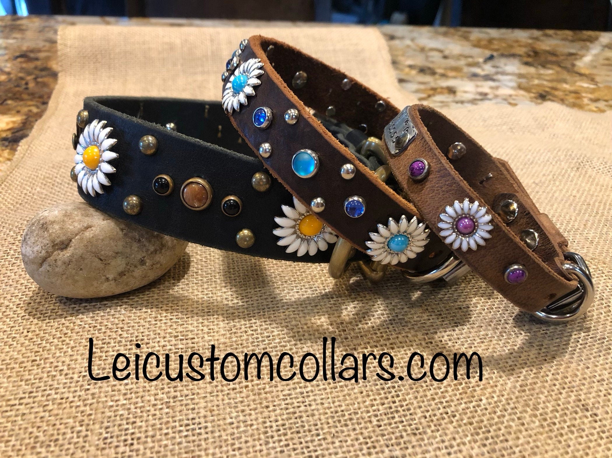 Handcrafted leather dog collars in black and brown tones featuring daisy concho accents, mixed metal studs, and colorful crystal details, styled on natural fabric and stone to showcase artisan variety and refined design